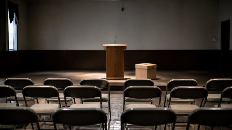 Rows of empty folding chairs facing a bare wooden lectern in a provincial town hall, harsh overhead lighting, cardboard ballot box nearby