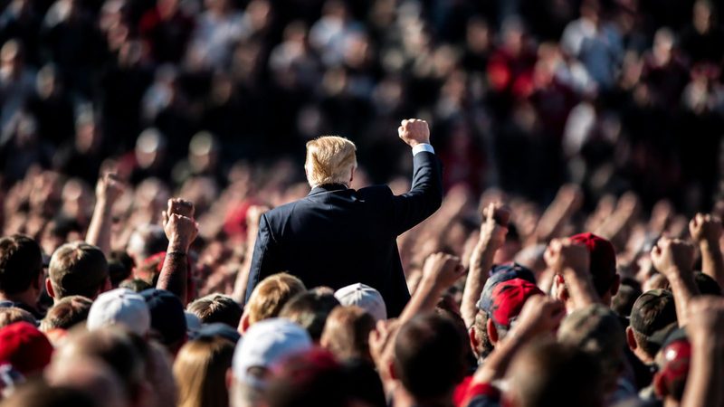 Crowded political rally seen from behind, hundreds of raised arms and hats, blurred faces packed tightly under bright afternoon sunlight